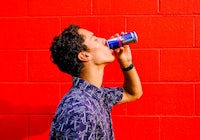 a man drinking a red bull can in front of a red brick wall